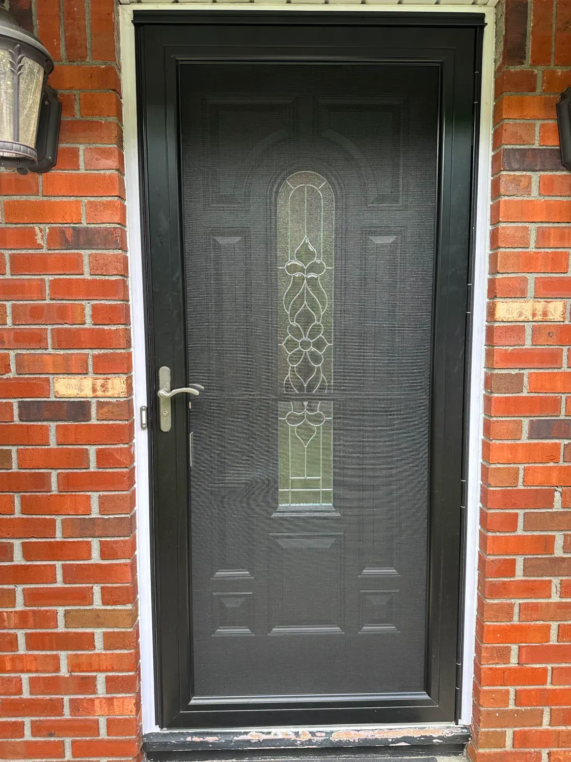 Black screen door with decorative glass panel, set in a brick exterior.