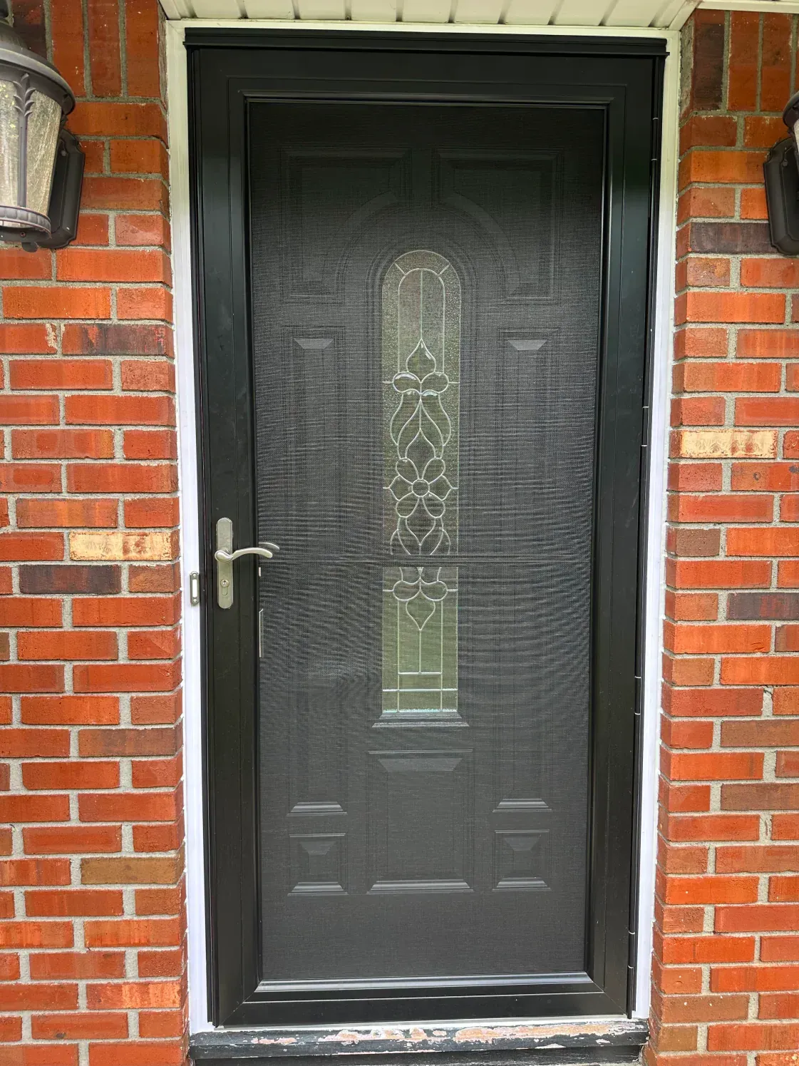 Black screen door with ornate glass panel, framed by a brick wall and white trim.