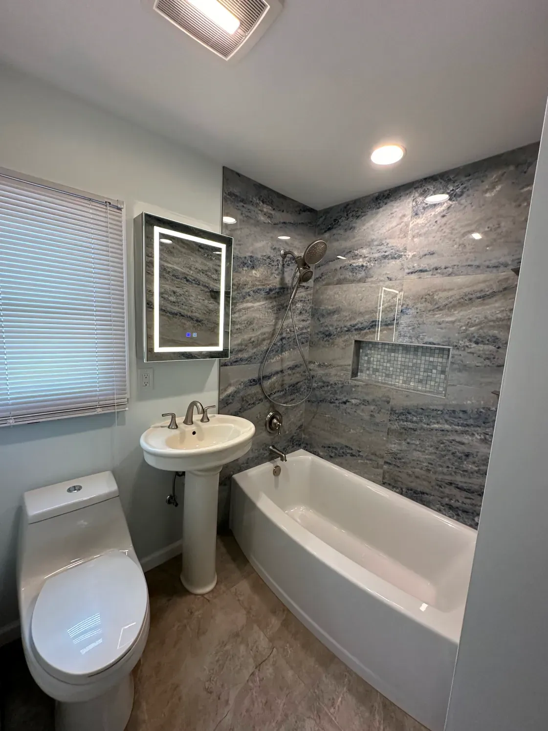 Modern bathroom with blue and gray tiled shower, white tub, toilet, pedestal sink, and lit mirror.