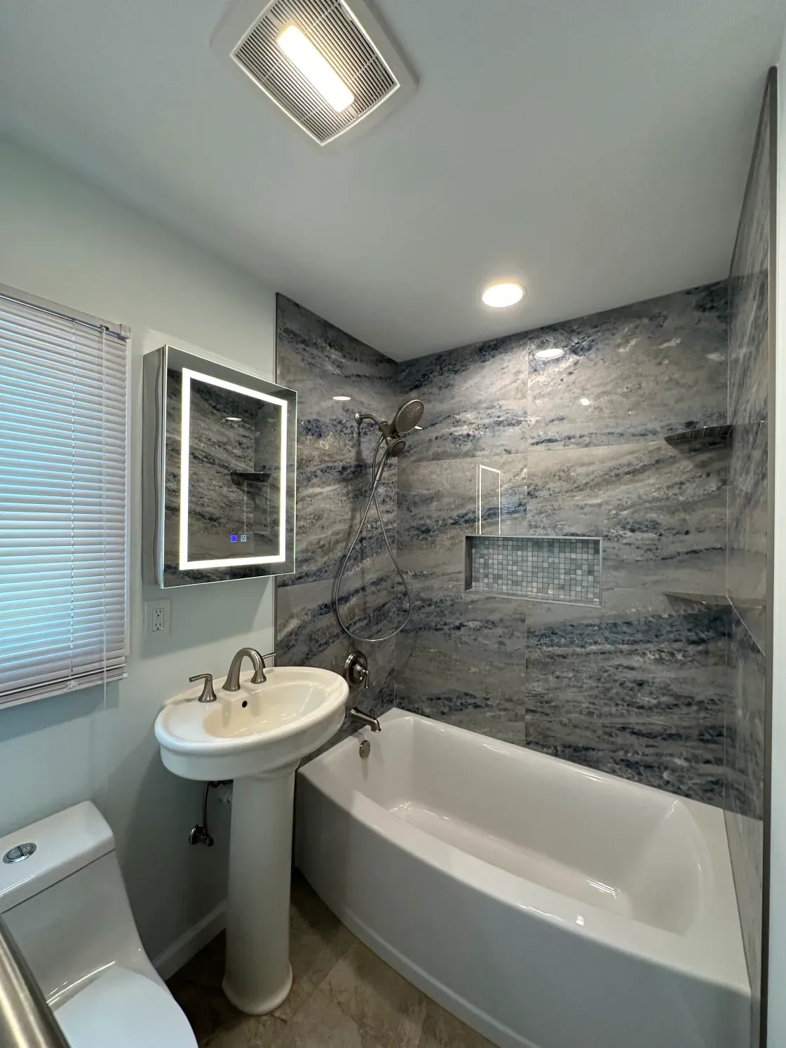 Bathroom with blue and gray patterned tile, white tub and sink, and a lit mirror.