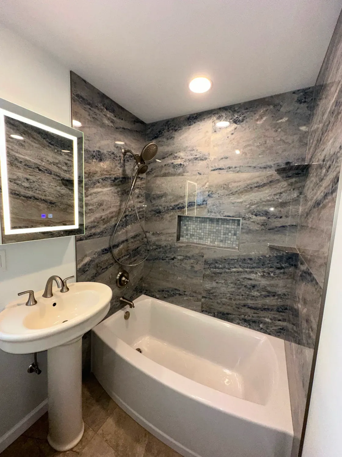 Bathroom with marble wall tiles, pedestal sink, bathtub, and lit mirror.