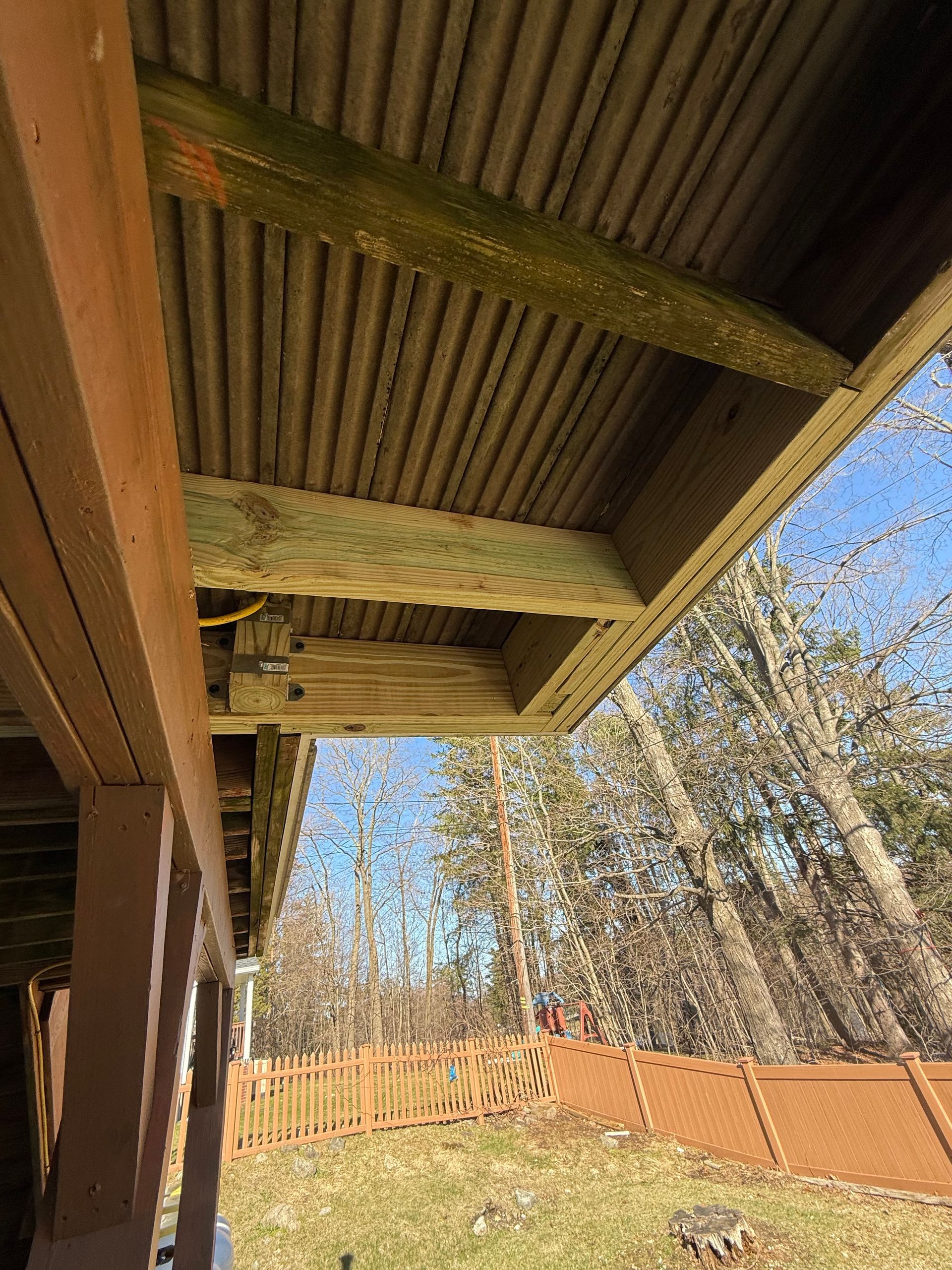 View from underneath a deck. Corrugated panels and wooden beams. Green moss on some wood. Brown fence in background.