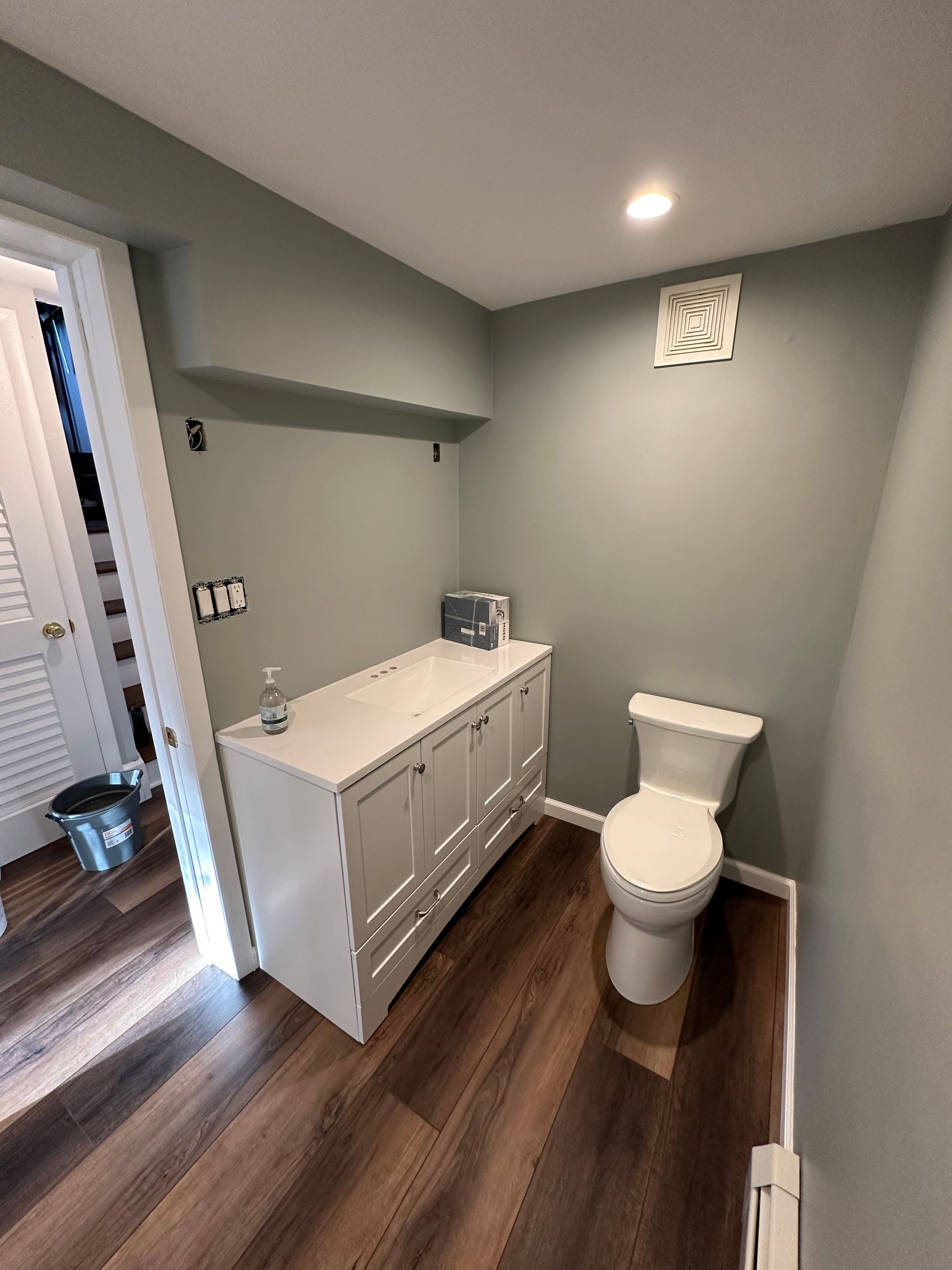 A small bathroom with a toilet, vanity, and open doorway, painted light green with wood flooring.