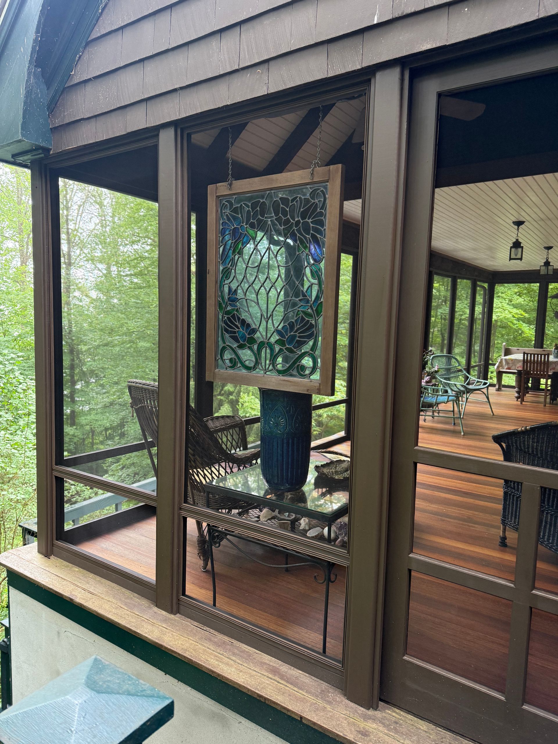 Screened porch with stained-glass art hanging on a blue vase, surrounded by lush green trees.