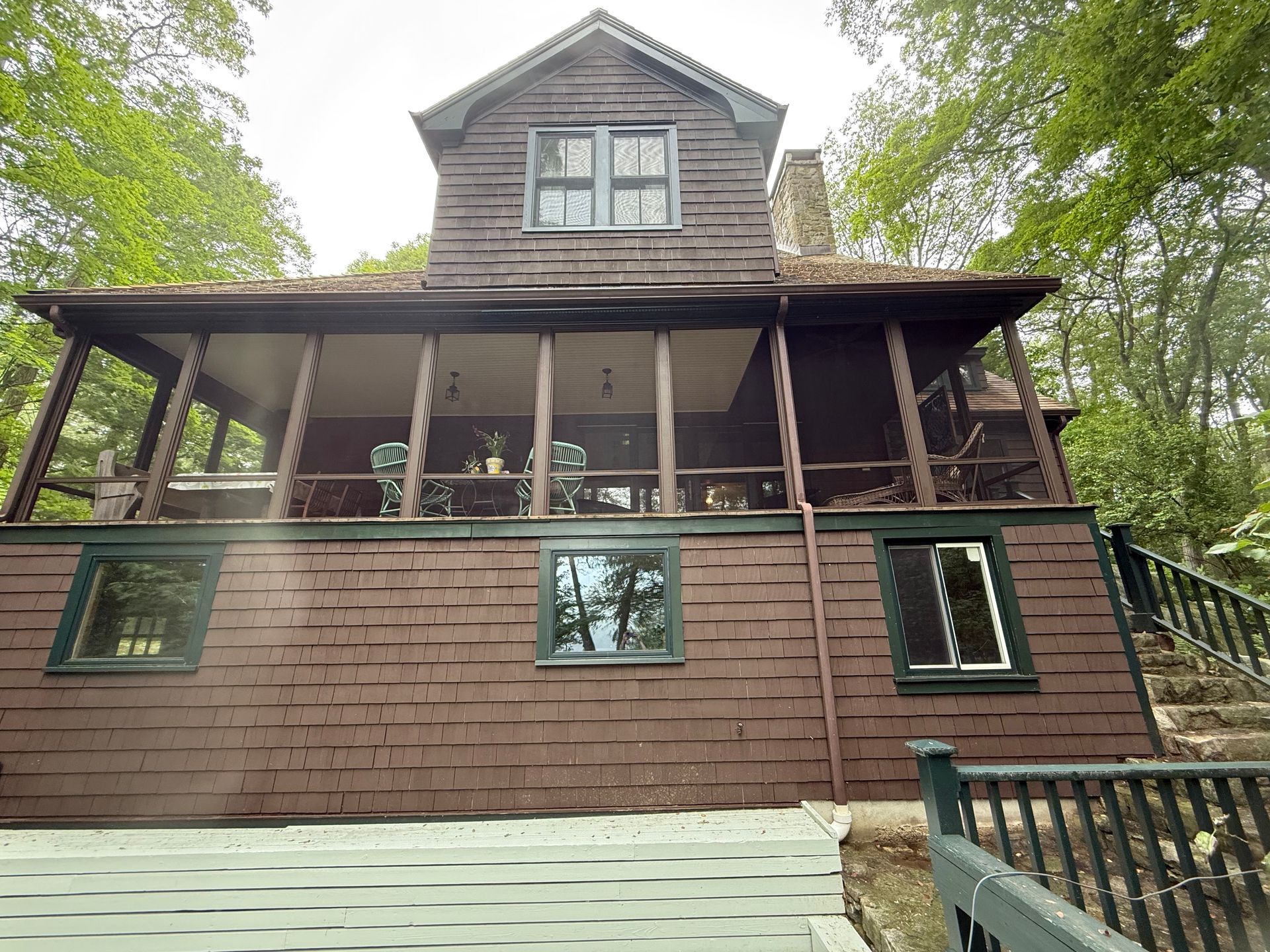 Brown, wooden house with a screened-in porch and windows. Nestled in a forest, overlooking a deck.