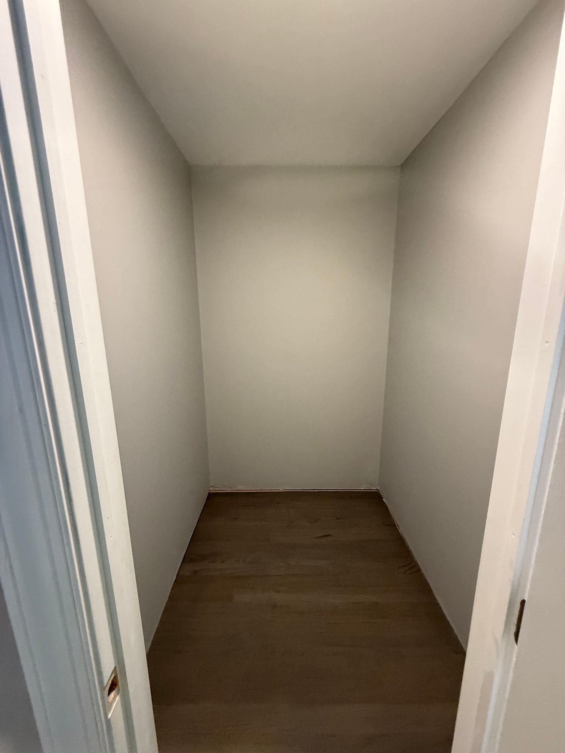 Empty, narrow closet with light gray walls, wooden floor, viewed from doorway.