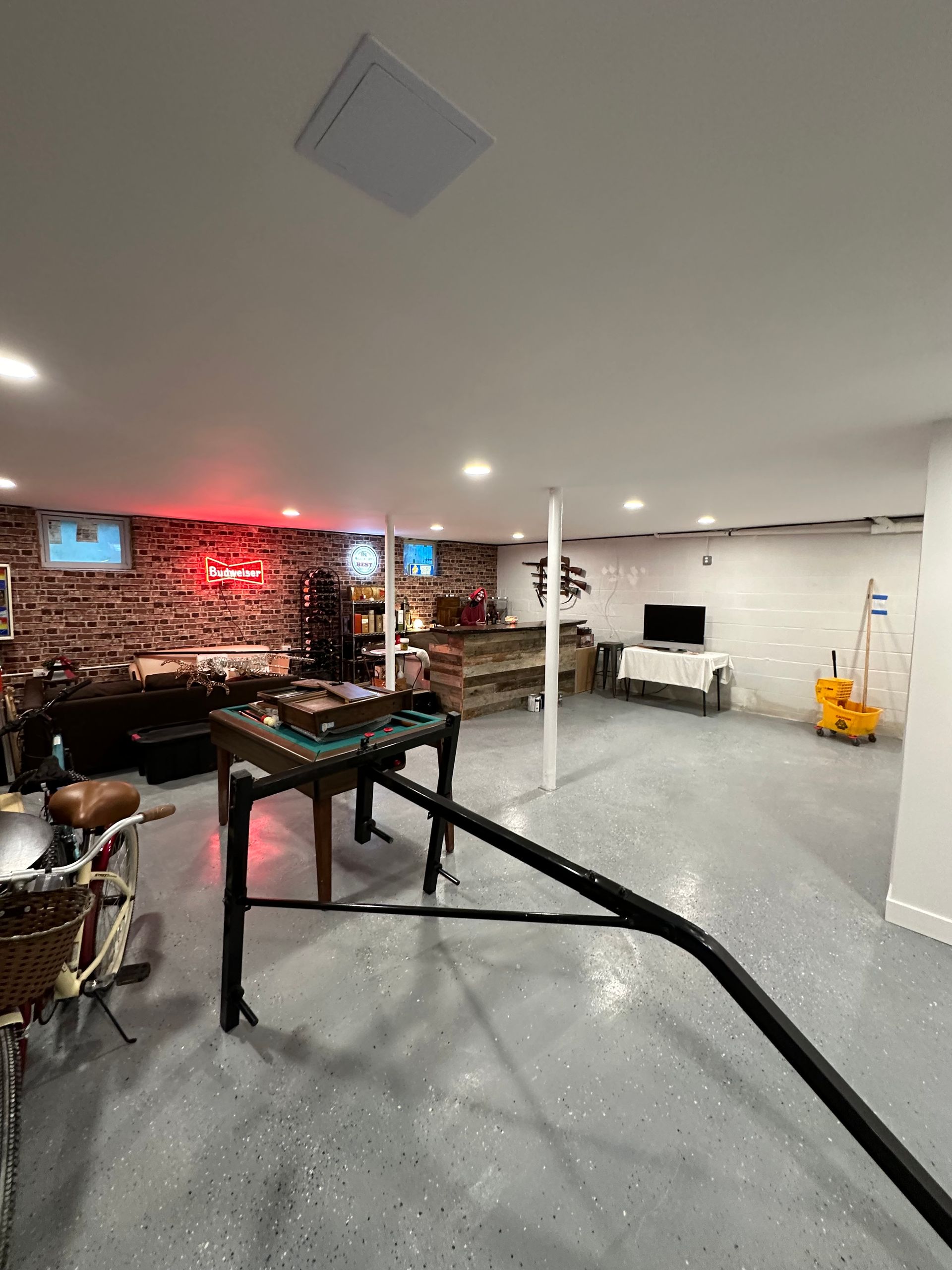 Basement with exposed brick wall, game table, TV, and polished gray floor. Fluorescent lighting brightens the space.