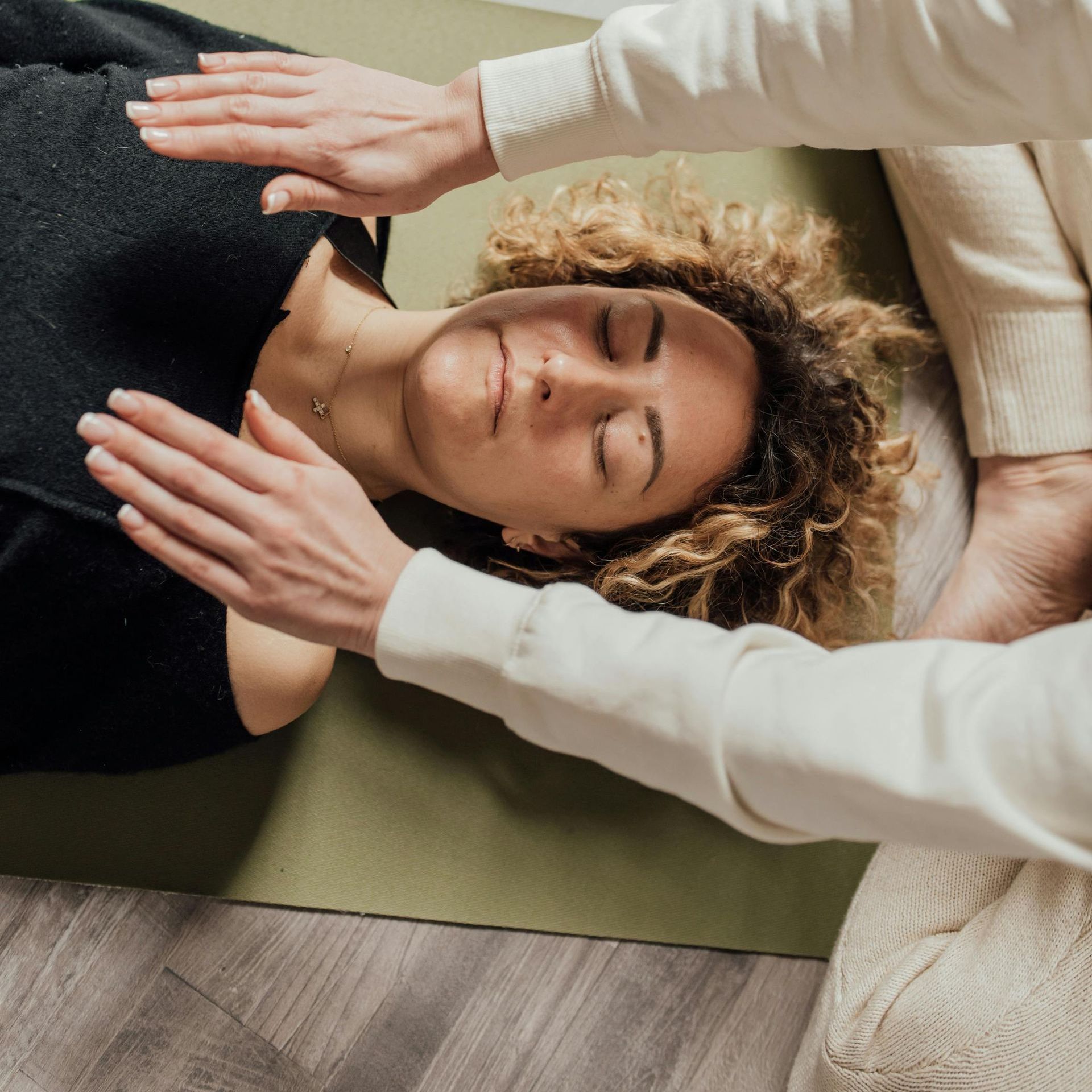 A woman is lying on a yoga mat with her eyes closed receiving Reiki.