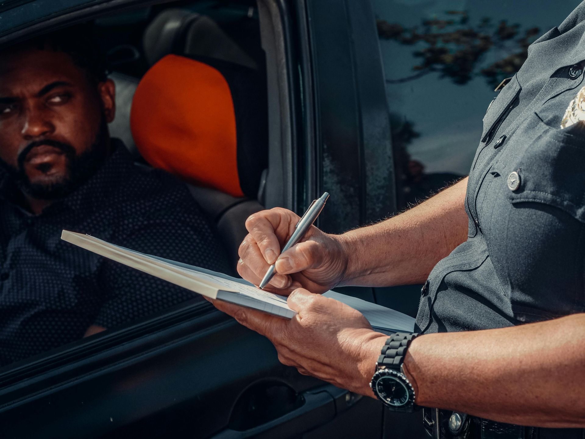 Police officer writing in a notebook next to a car. A man in the car looks on.