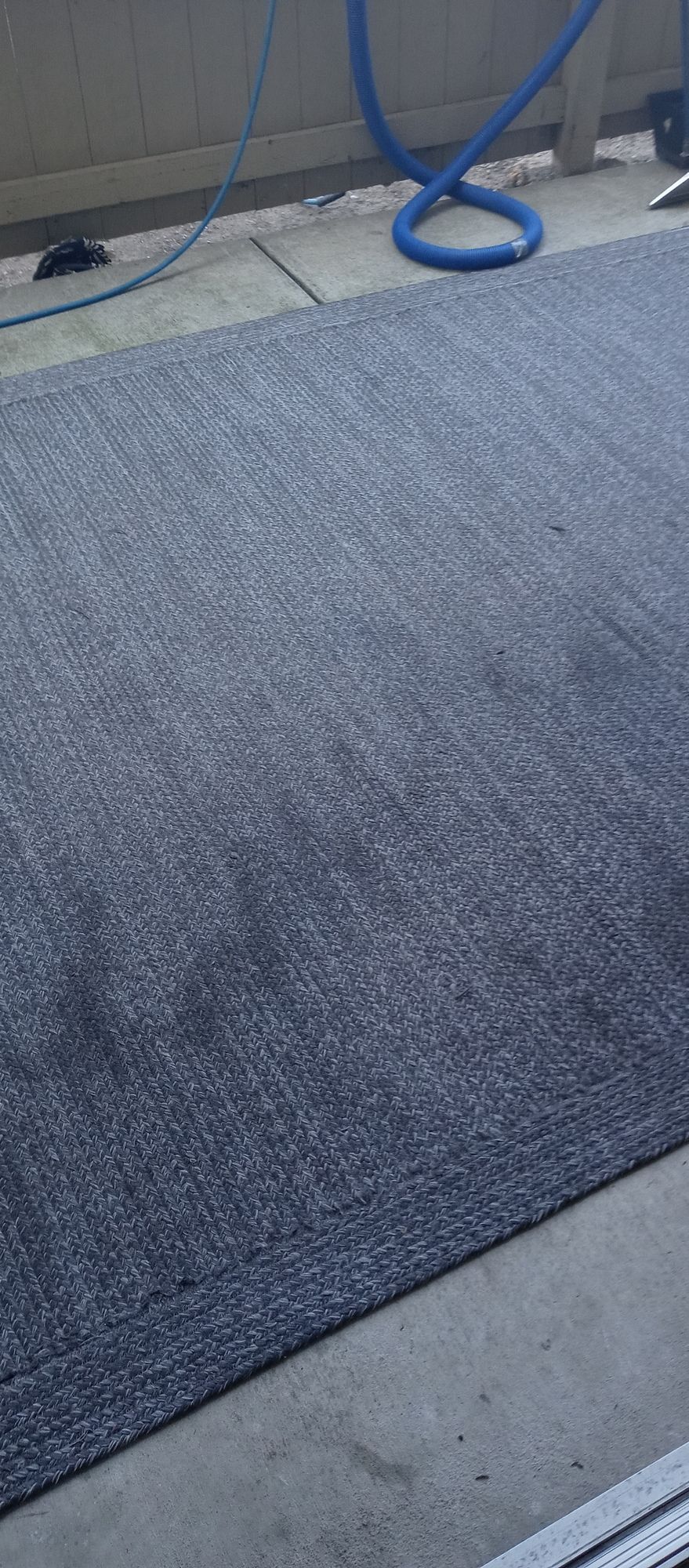 A gray rug is sitting on a concrete floor next to a blue hose.