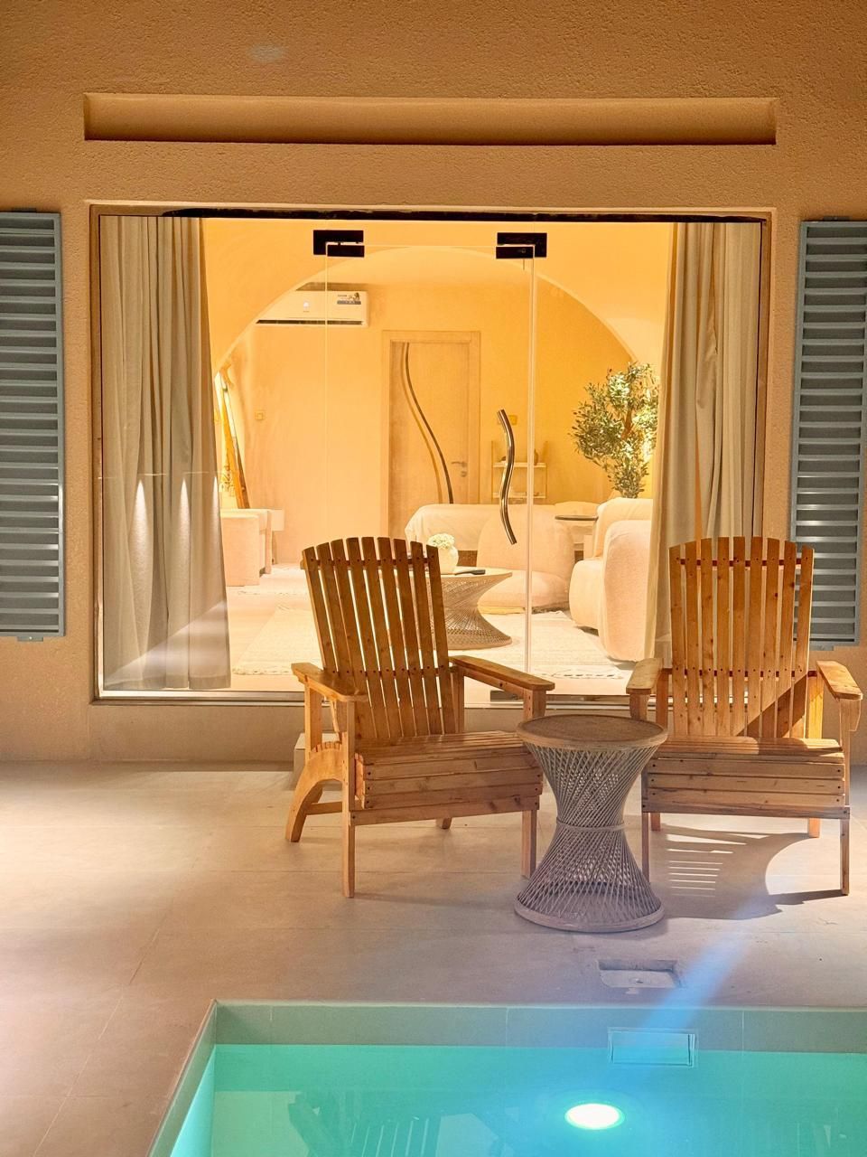 Outdoor patio with wooden chairs and small table, overlooking a light-filled interior through large glass doors.