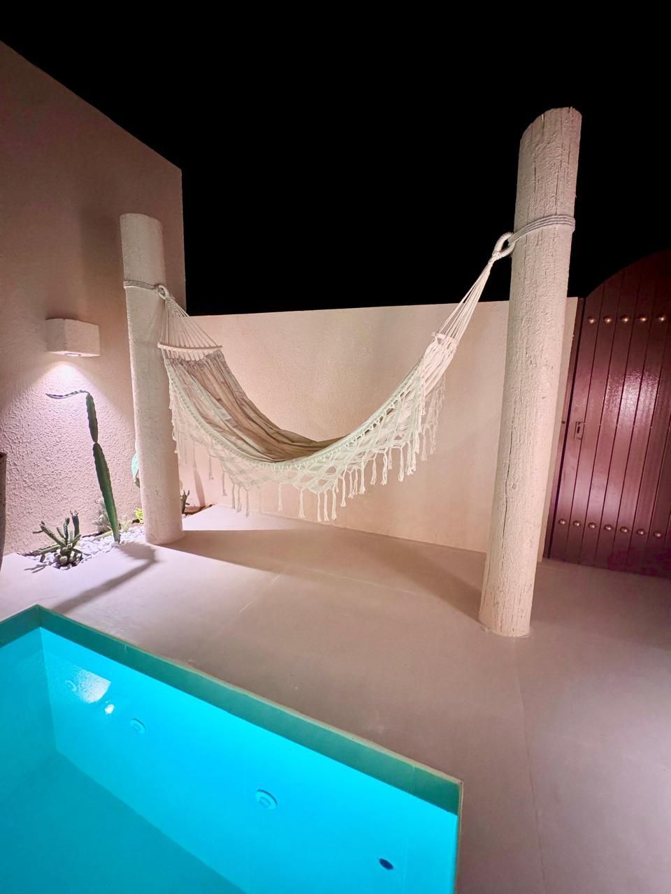 Outdoor scene at night: white hammock between two posts, small pool with blue water, cactus, and wall with dim light.