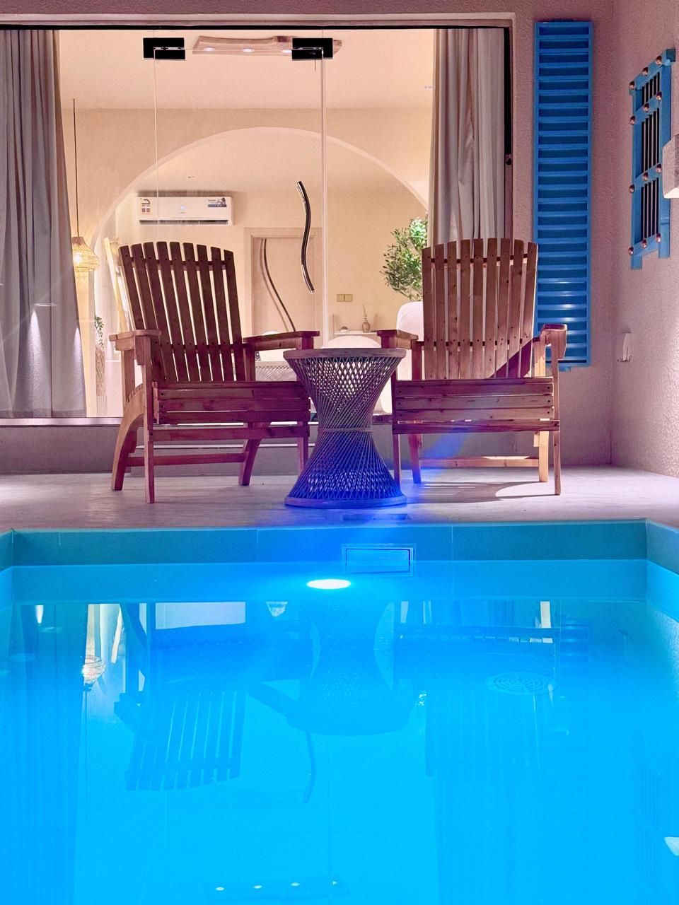 Pool with blue water; wooden chairs and small table on patio in front of a window. Blue shutter on right.