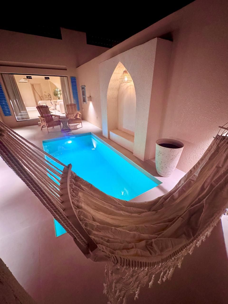 Cozy, enclosed pool area with a hammock, archway, and lit pool. Beige walls, night setting.