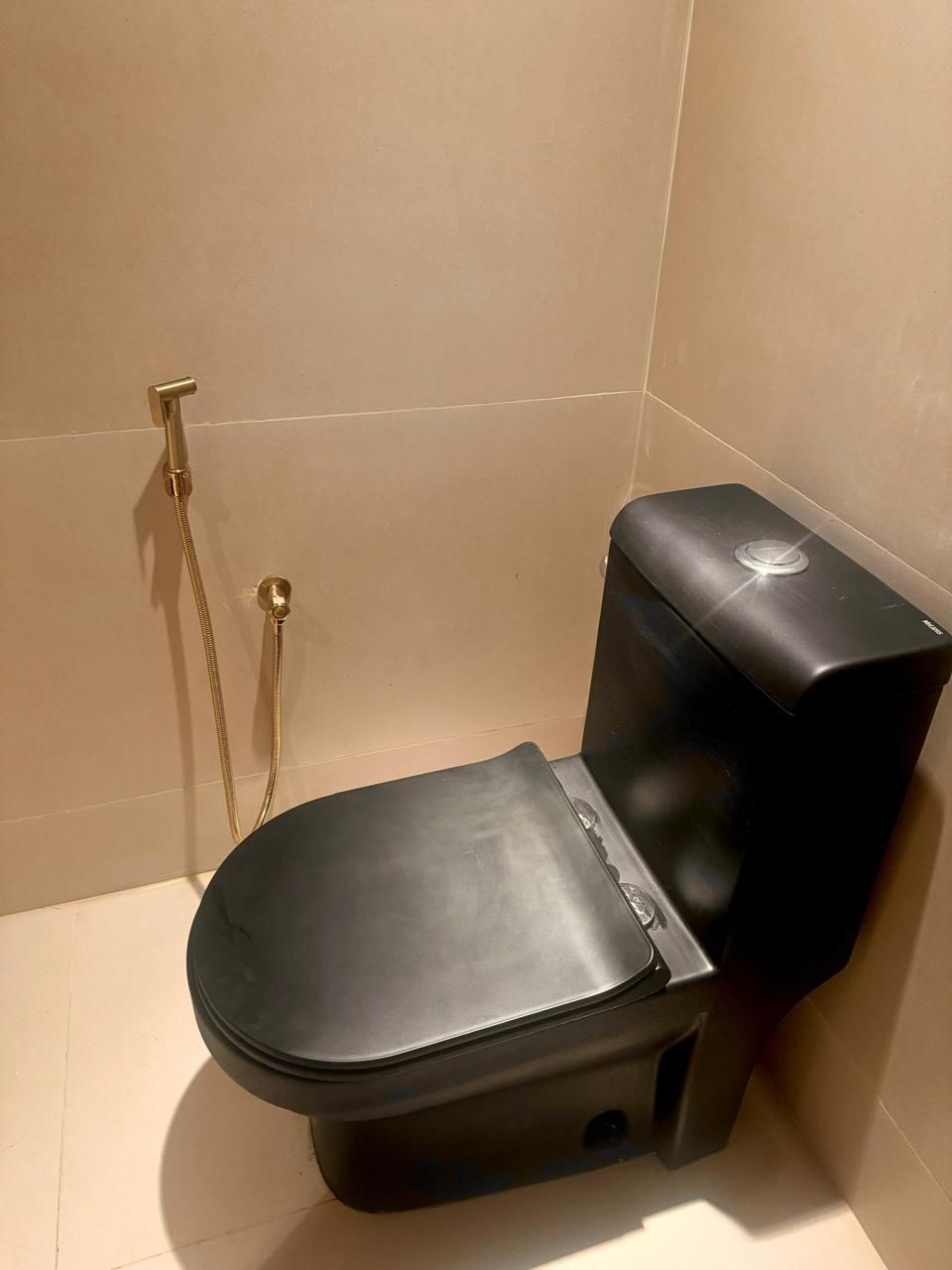 Black toilet in a corner with a gold bidet sprayer on a neutral tiled wall.