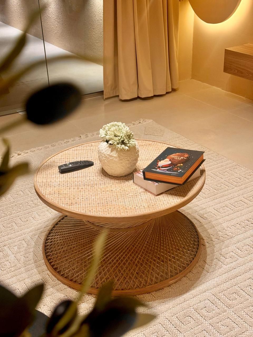 Round rattan coffee table with books, vase of flowers, and a remote on a rug.