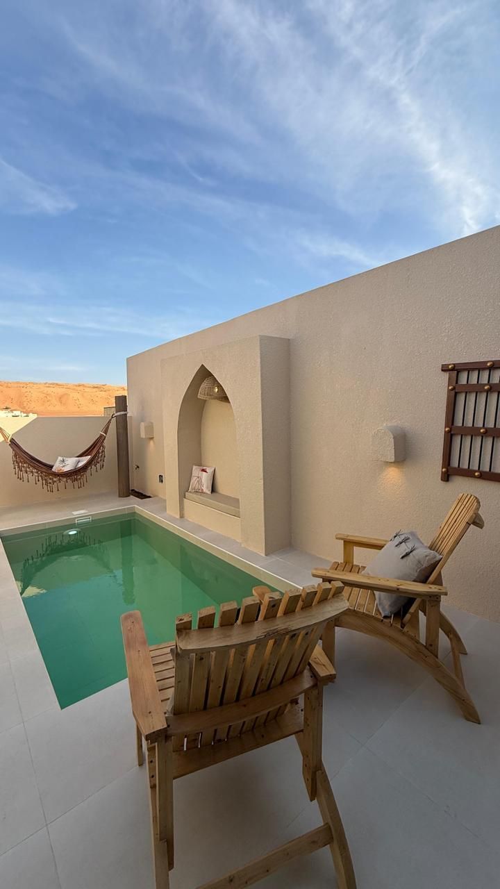 Private pool area with lounge chairs and hammock, against a beige wall, under a blue sky.