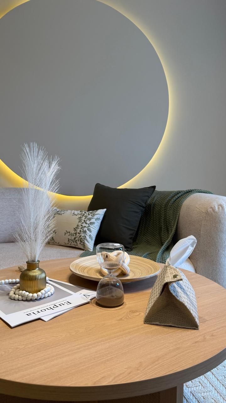 Cozy living space with a round wooden table, sofa with pillows, and a large circular wall feature with warm lighting.