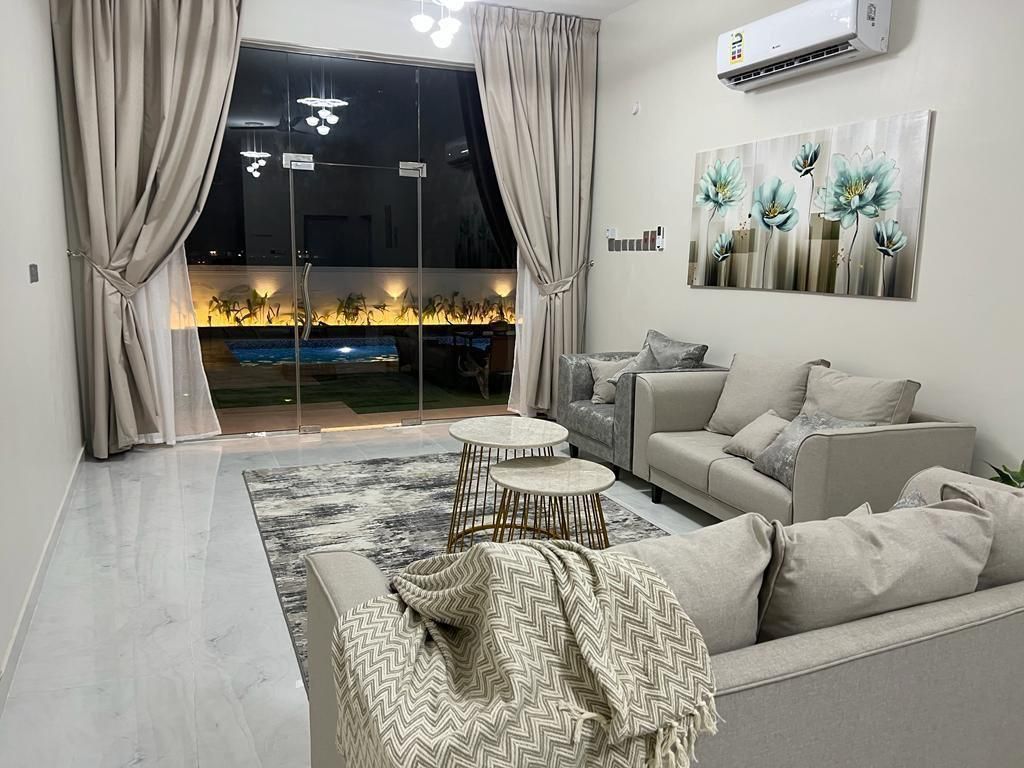 Living room with beige couches, rug, curtains, glass wall overlooking a pool, artwork, and coffee tables.