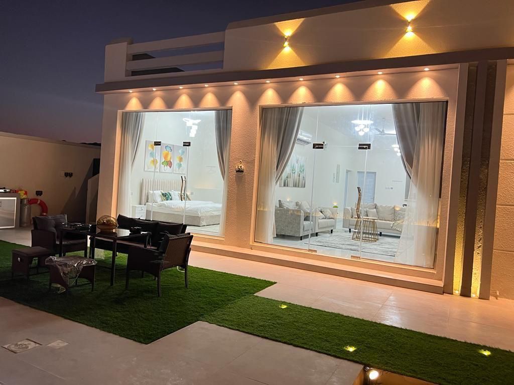 Exterior view of a lit modern building with glass doors revealing a white interior, outdoor seating on the grass.