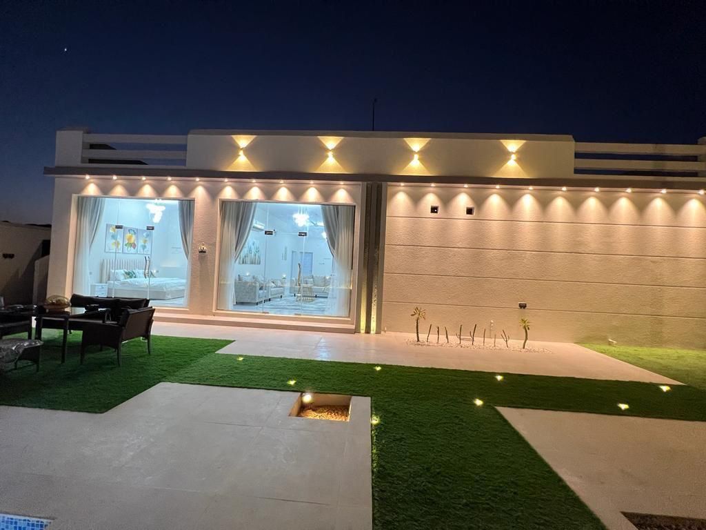 Exterior of a lit-up white building at night, with an illuminated interior visible through doorways. Artificial lawn.