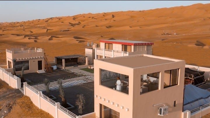 Desert houses with a tan exterior and red roof, surrounded by sand dunes and a white wall.