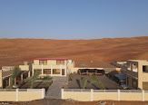 Buildings with light-colored walls and red roofs in a desert landscape with reddish sand dunes in the background.