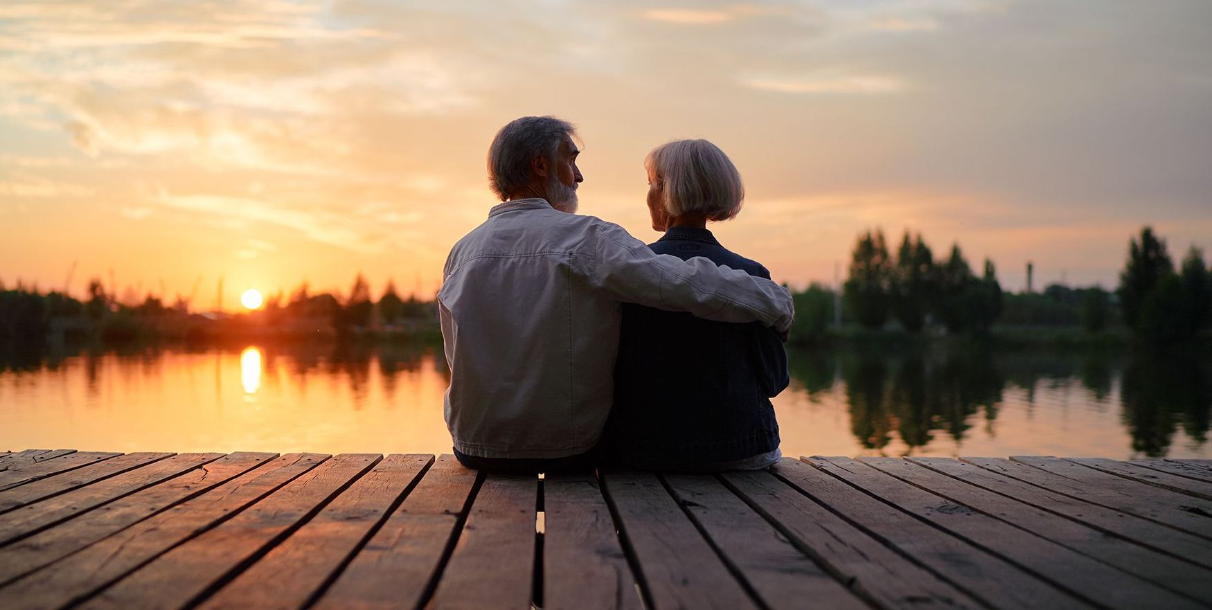 An elderly couple is sitting on a dock watching the sunset over a lake.