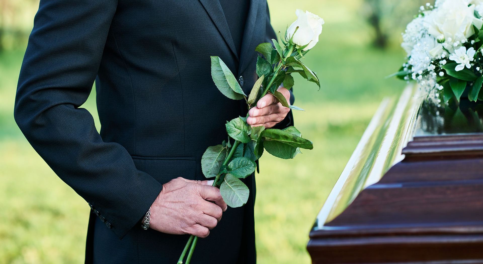 A man in a suit is holding a white rose in front of a coffin.