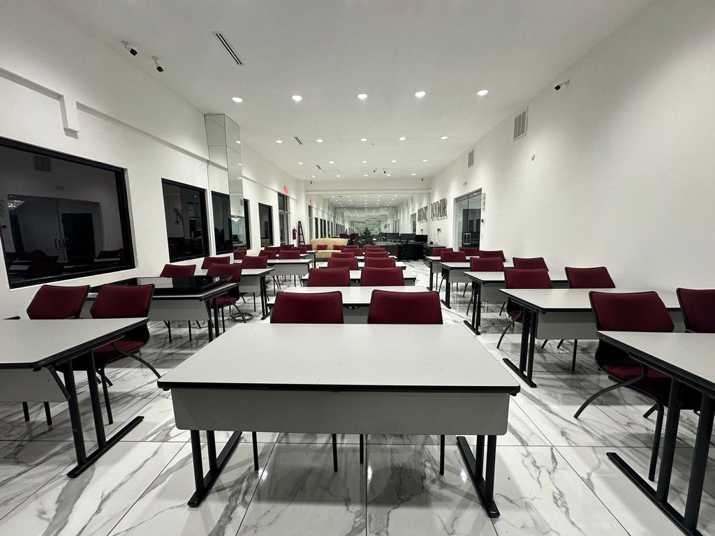 Empty classroom with rows of tables and chairs, white walls, and large windows.