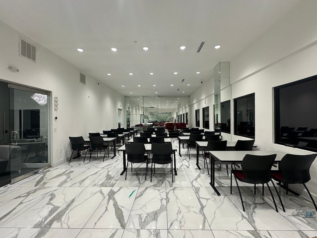 Bright, open room with tables and chairs, likely a cafeteria or dining area. Large windows, mirrored columns, and marble-look floor.