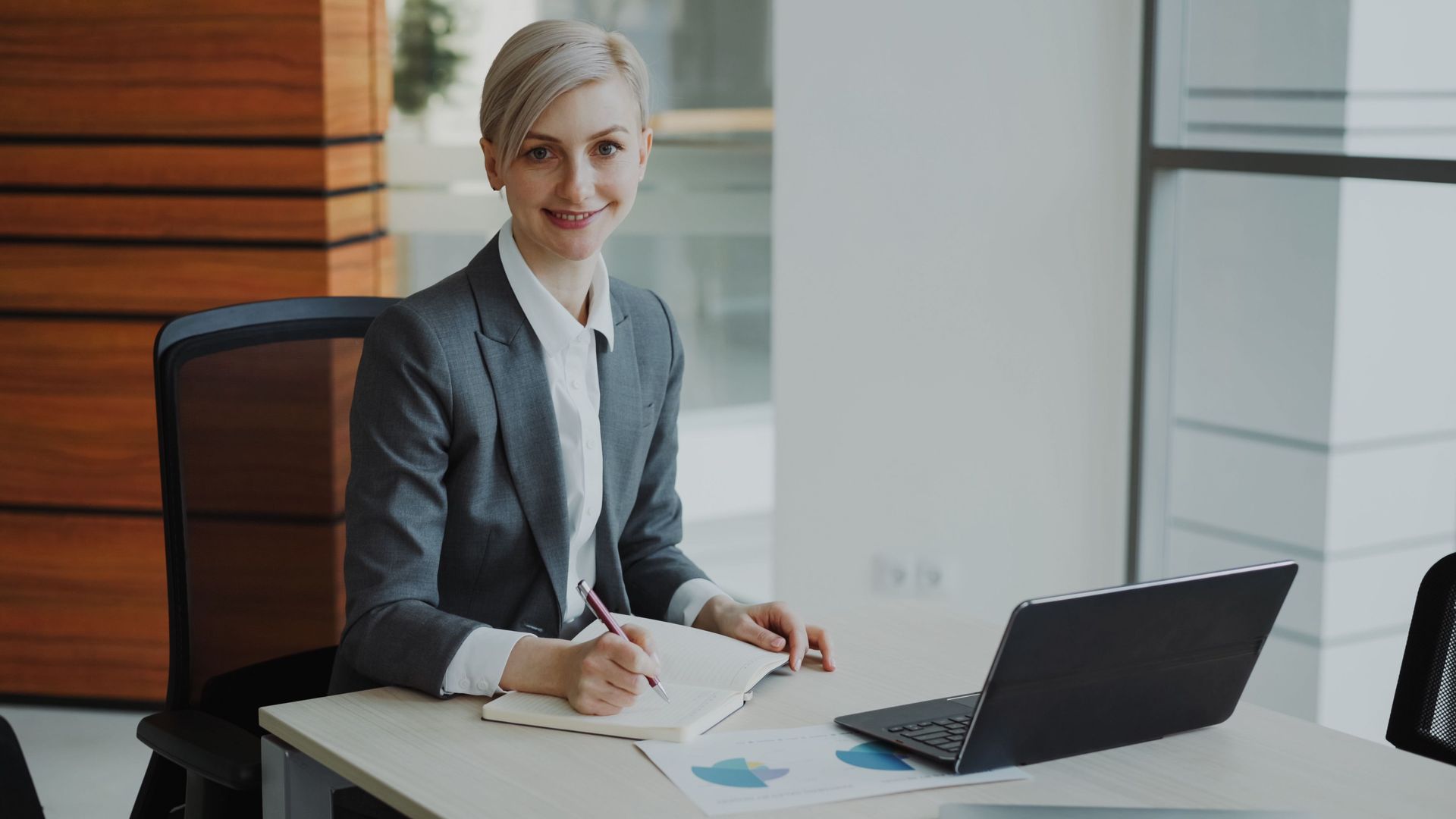 Person seated at a desk, smiling and writing beside an open laptop in a bright office.