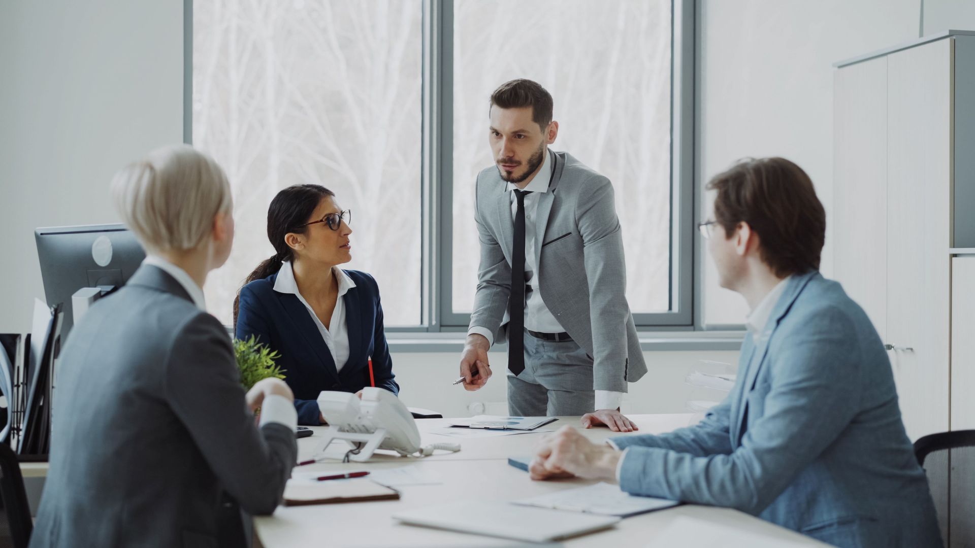 Business team in a bright meeting room, with one man presenting to three seated colleagues.
