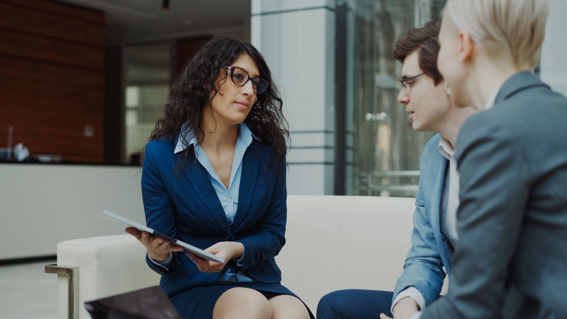 Three coworkers in business attire discuss on a sofa in a modern office lobby