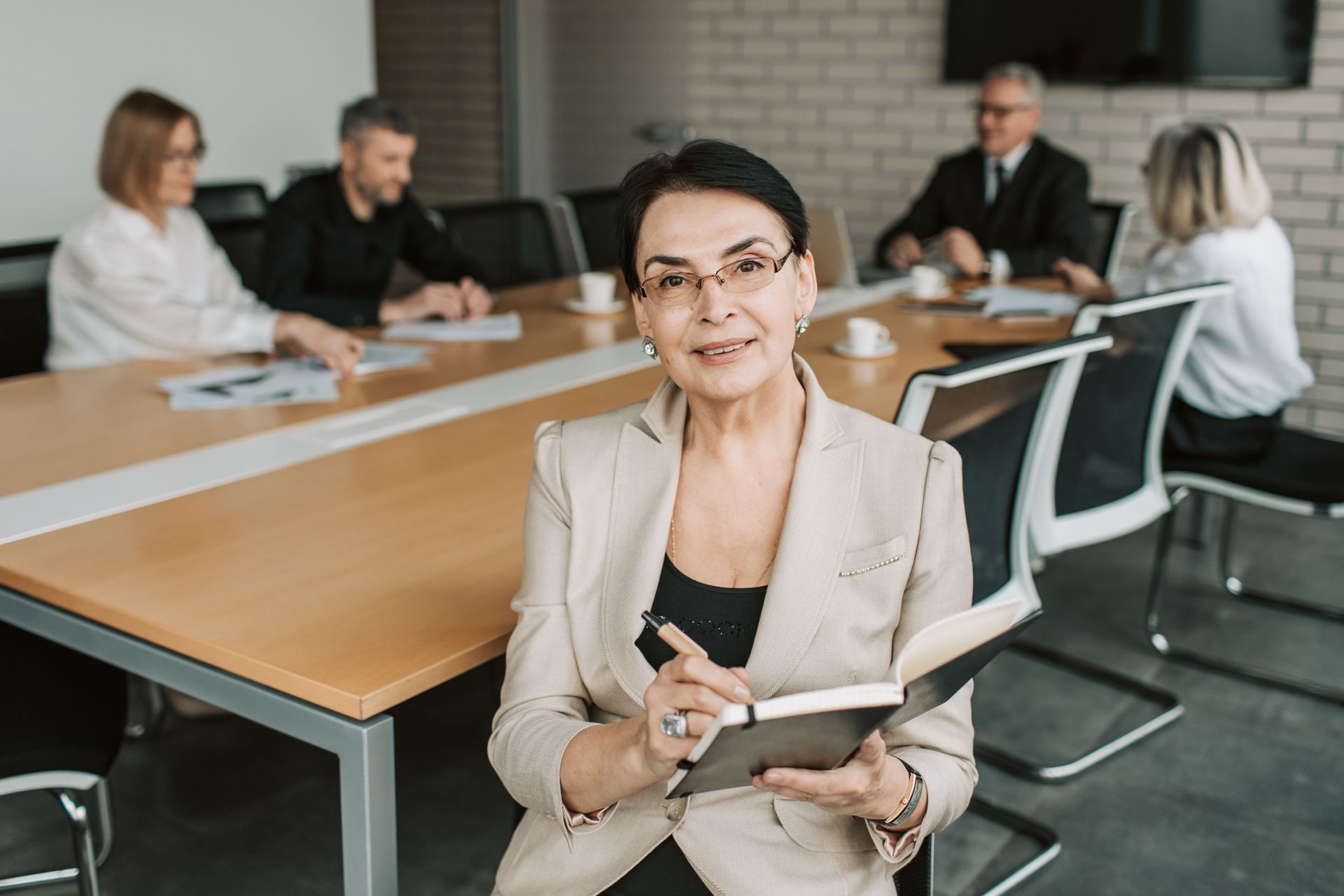 Woman holding a notebook in a modern conference room with colleagues seated around a table