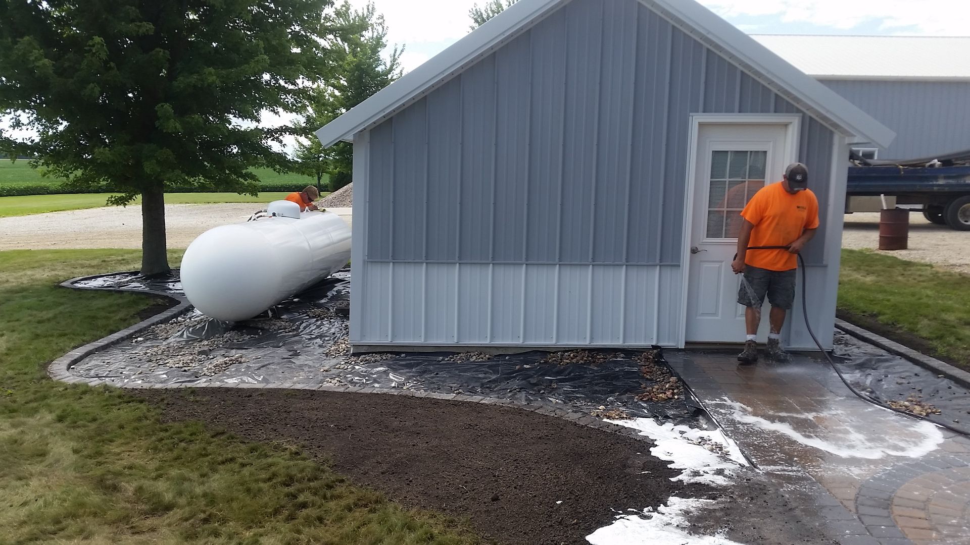 A person in an orange shirt uses a pressure washer to clean a walkway leading to a gray outbuilding near a propane tank.