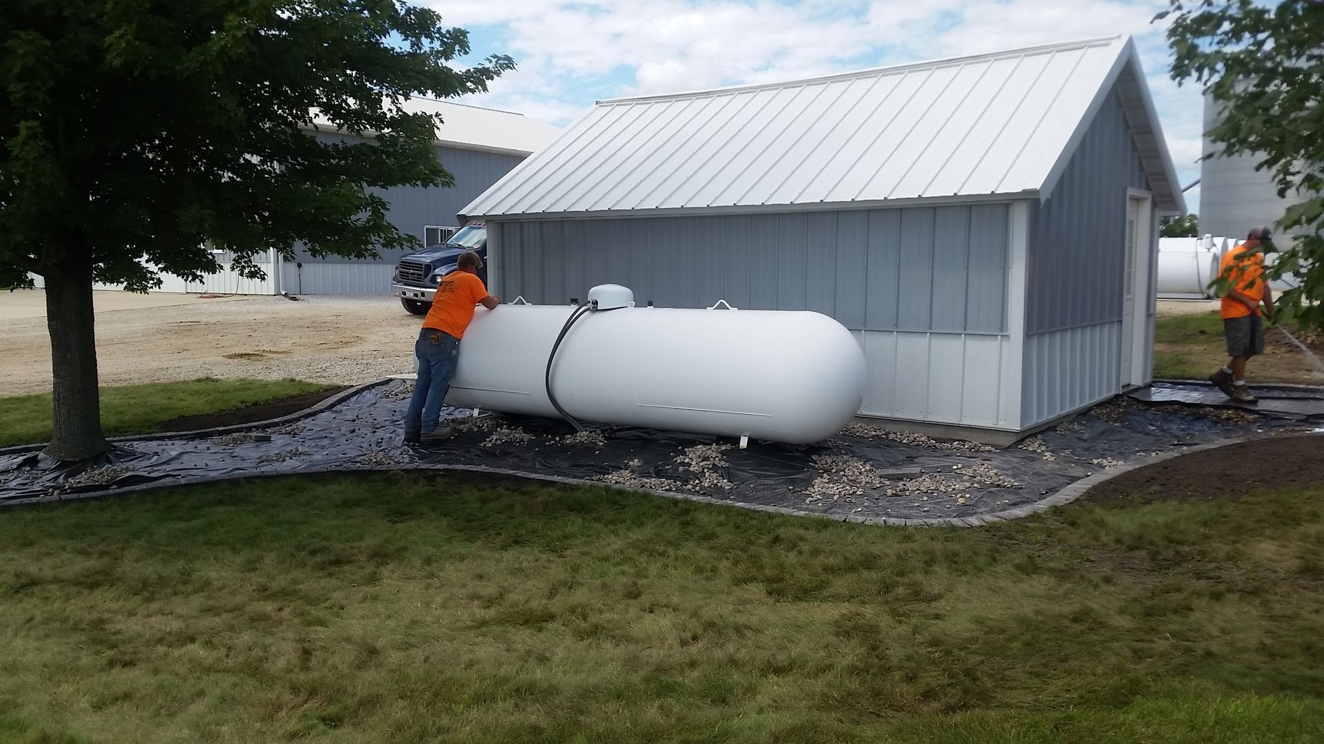 Two workers in orange shirts install a white propane tank next to a small building on a gravel and grass lot.