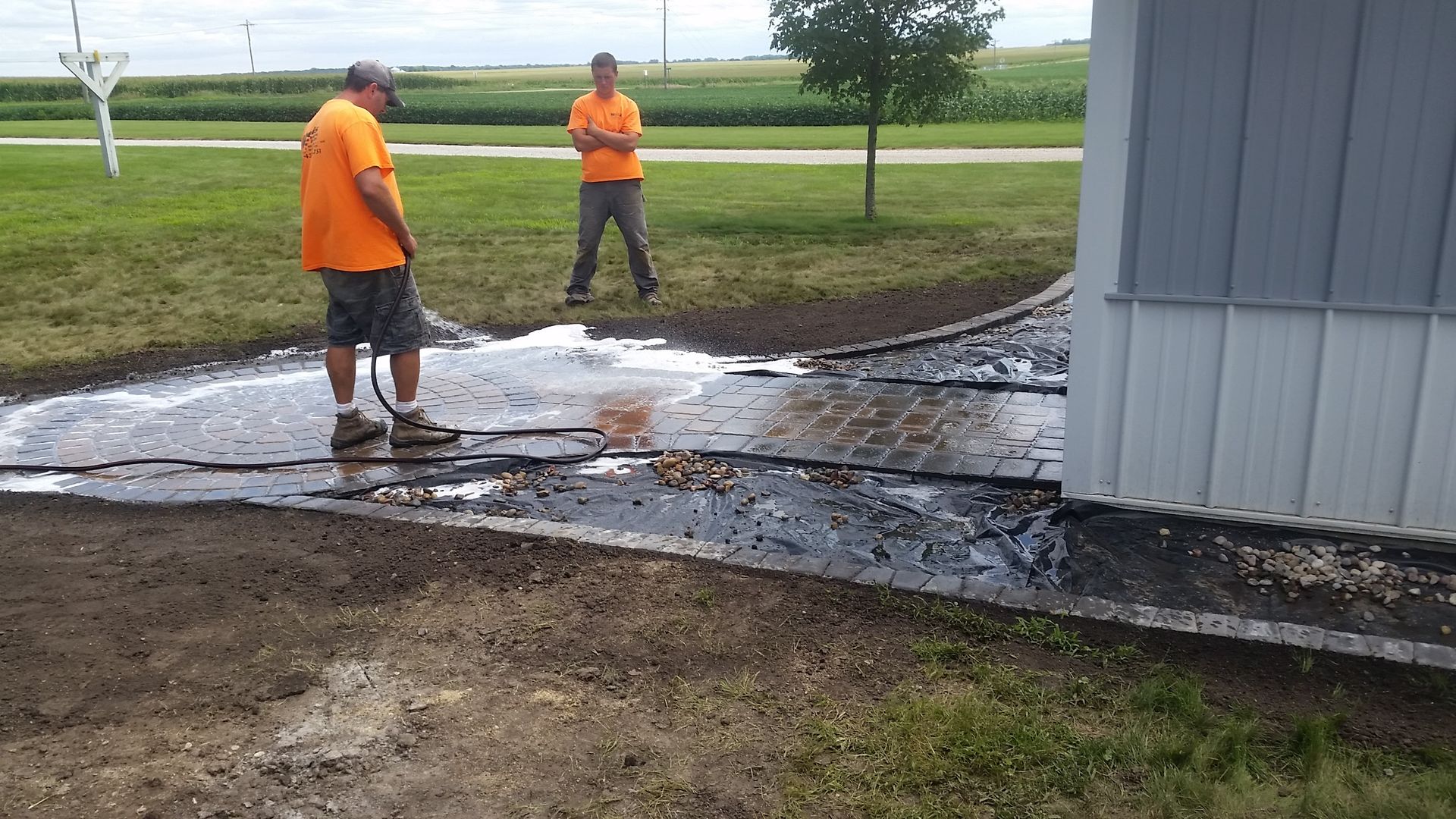 Two people in orange shirts work on a flooded paved area outside a building in a rural setting.
