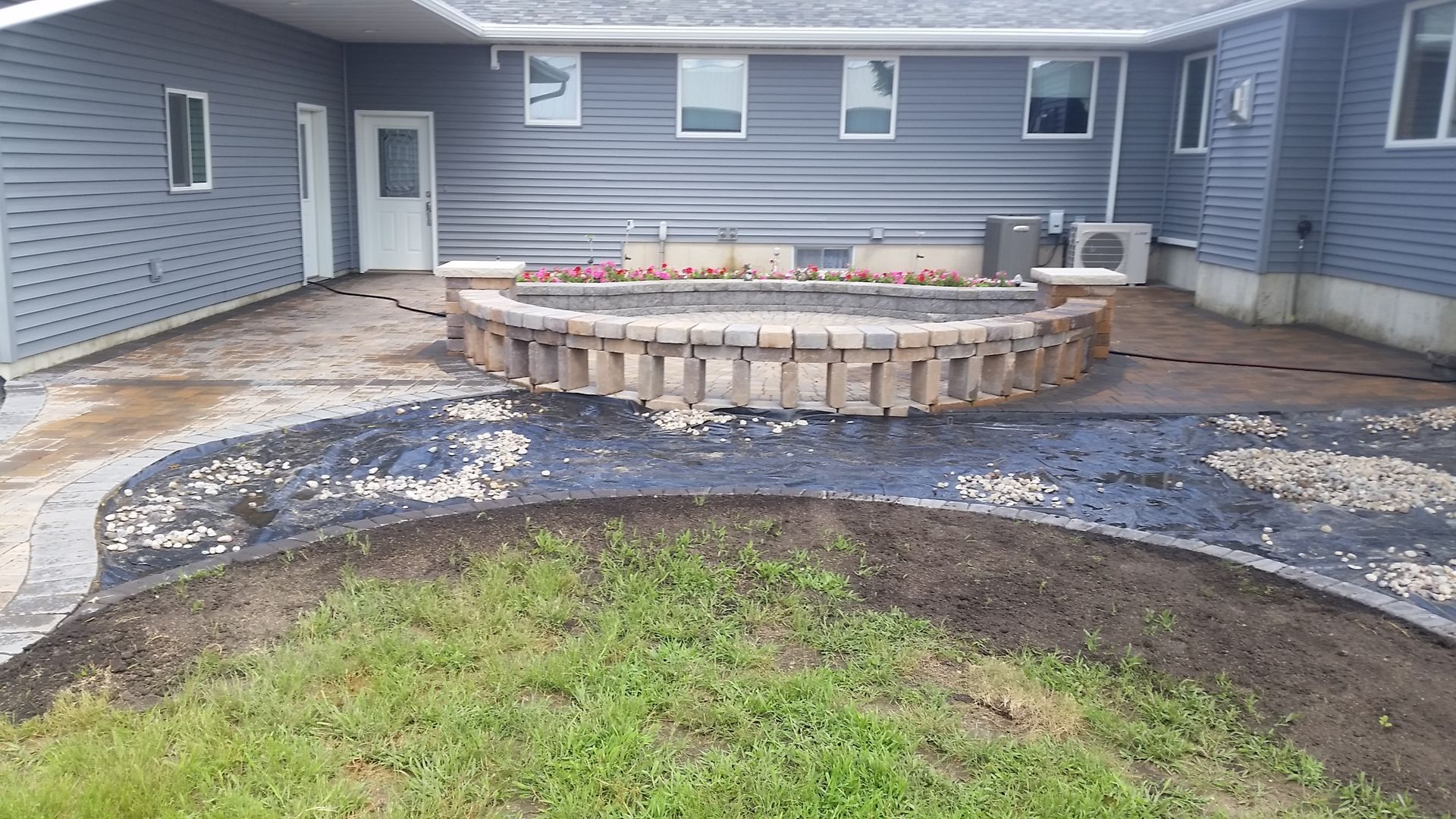 A gray house with a curved stone retaining wall patio, landscaped border, and gravel area under construction.