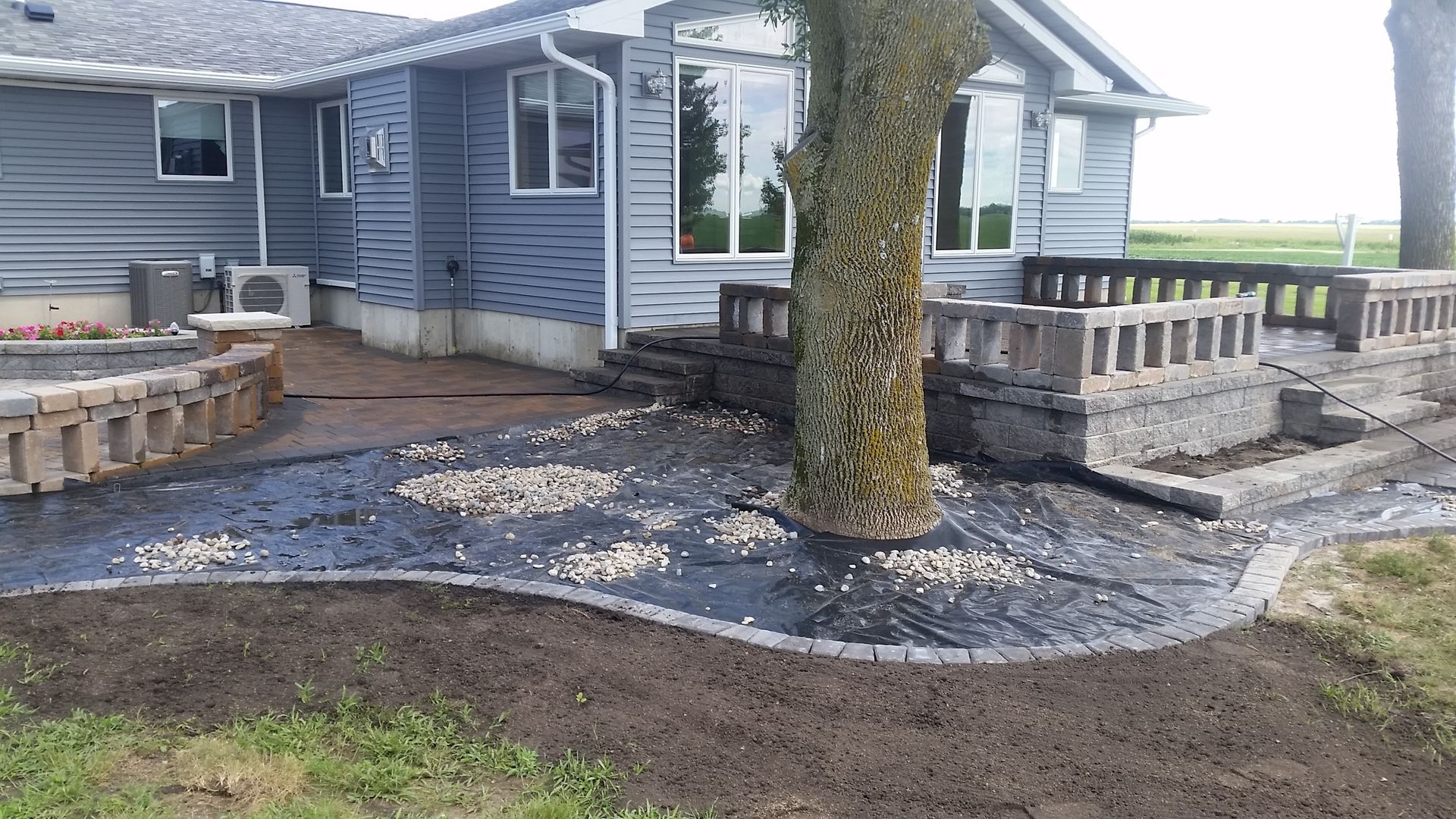A gray house exterior with a landscaped patio area featuring stone retaining walls, landscape fabric, and a large tree.