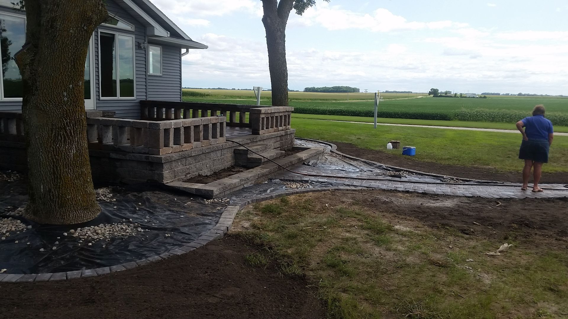 A person stands by a house with a stone porch and a garden pathway undergoing landscaping in a rural backyard.