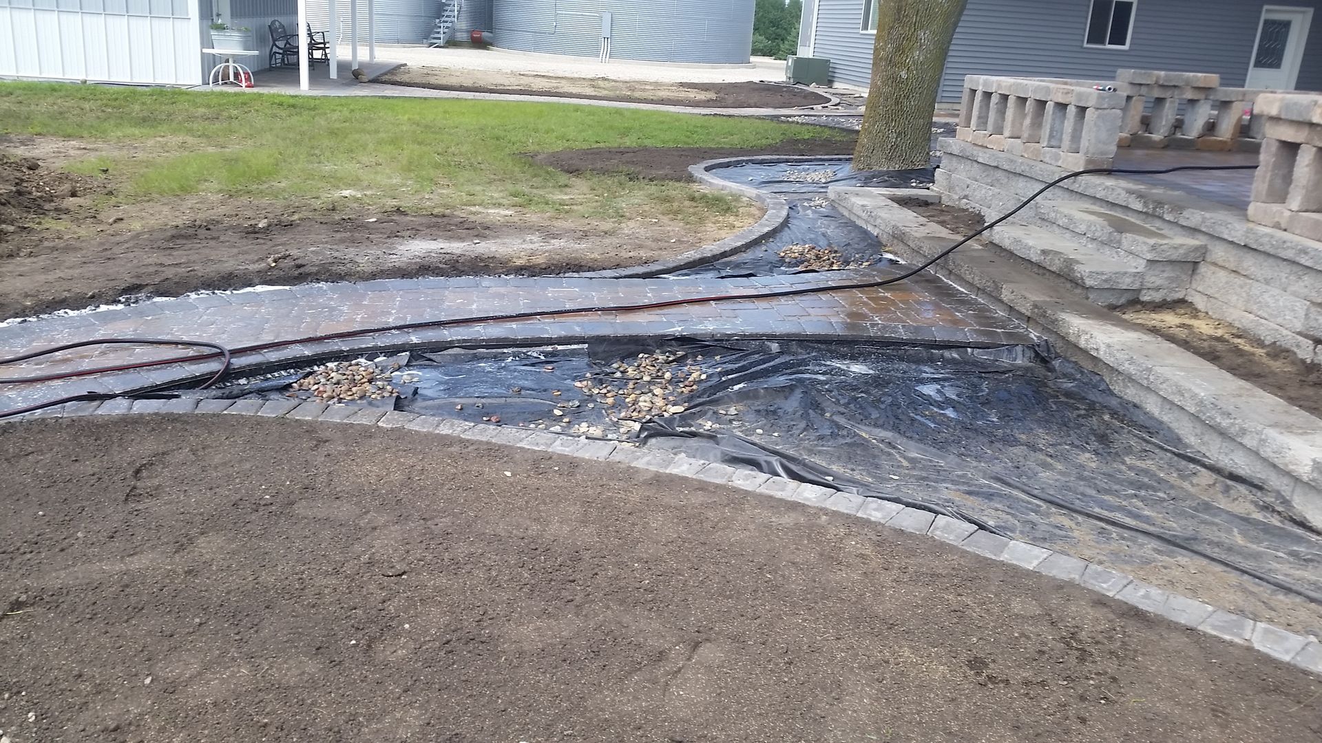 An outdoor patio area under construction, featuring a curved paver border, gravel base, and landscape fabric near a porch.