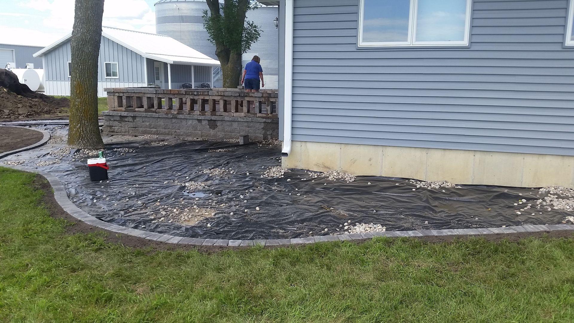A person works behind a concrete block wall near a house foundation with a newly defined landscape bed filled with mulch.