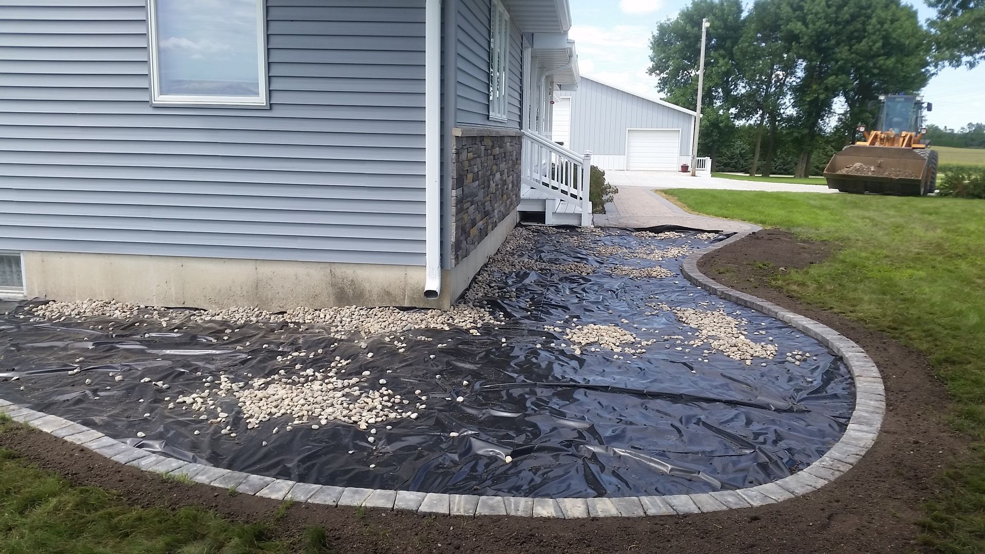 Landscaped area next to a house with a black ground cover sheet, a stone border, and scattered decorative rocks.