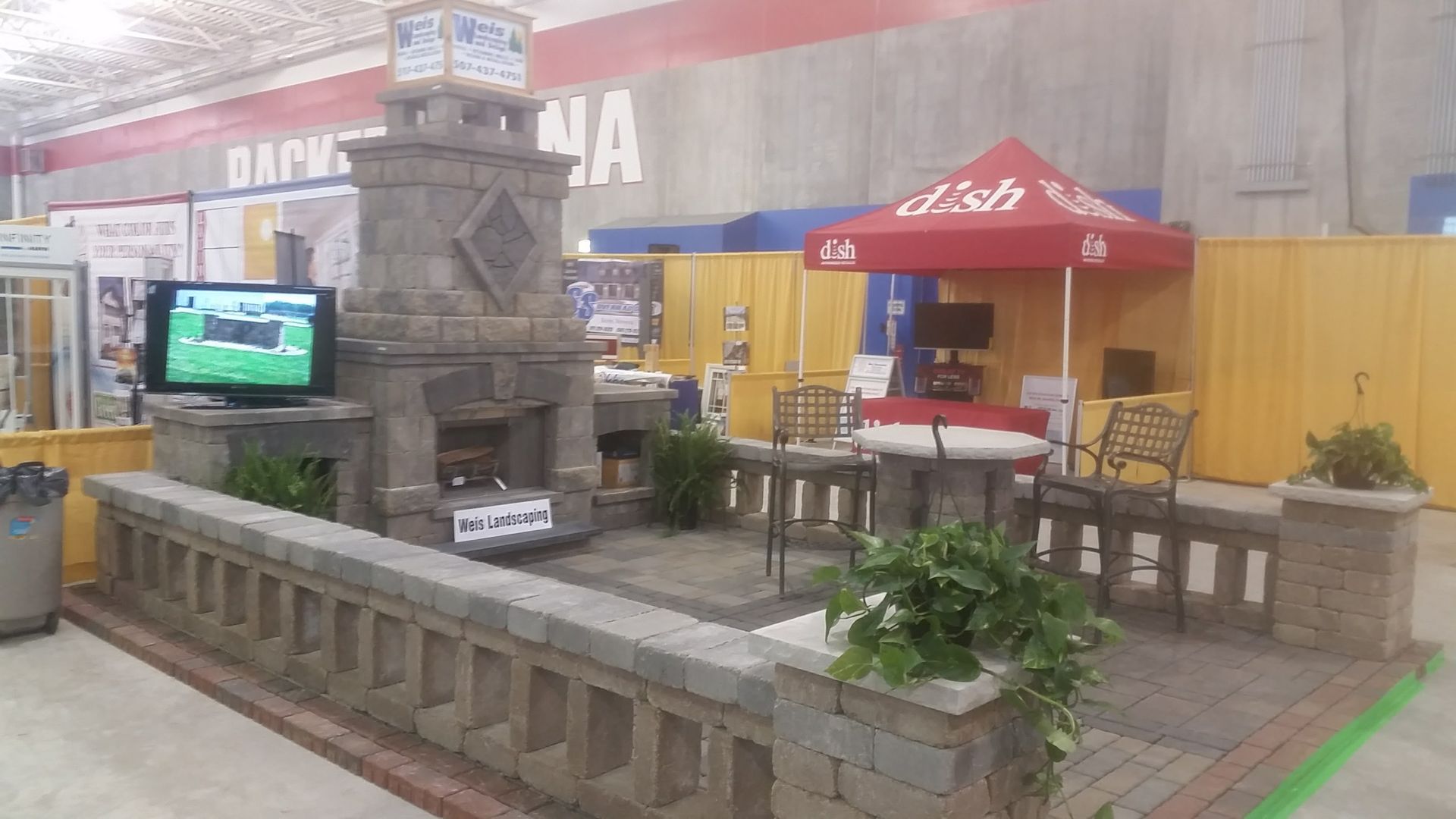 A display booth features a stone fireplace, patio furniture, and a Dish-branded tent at an indoor exhibition hall.