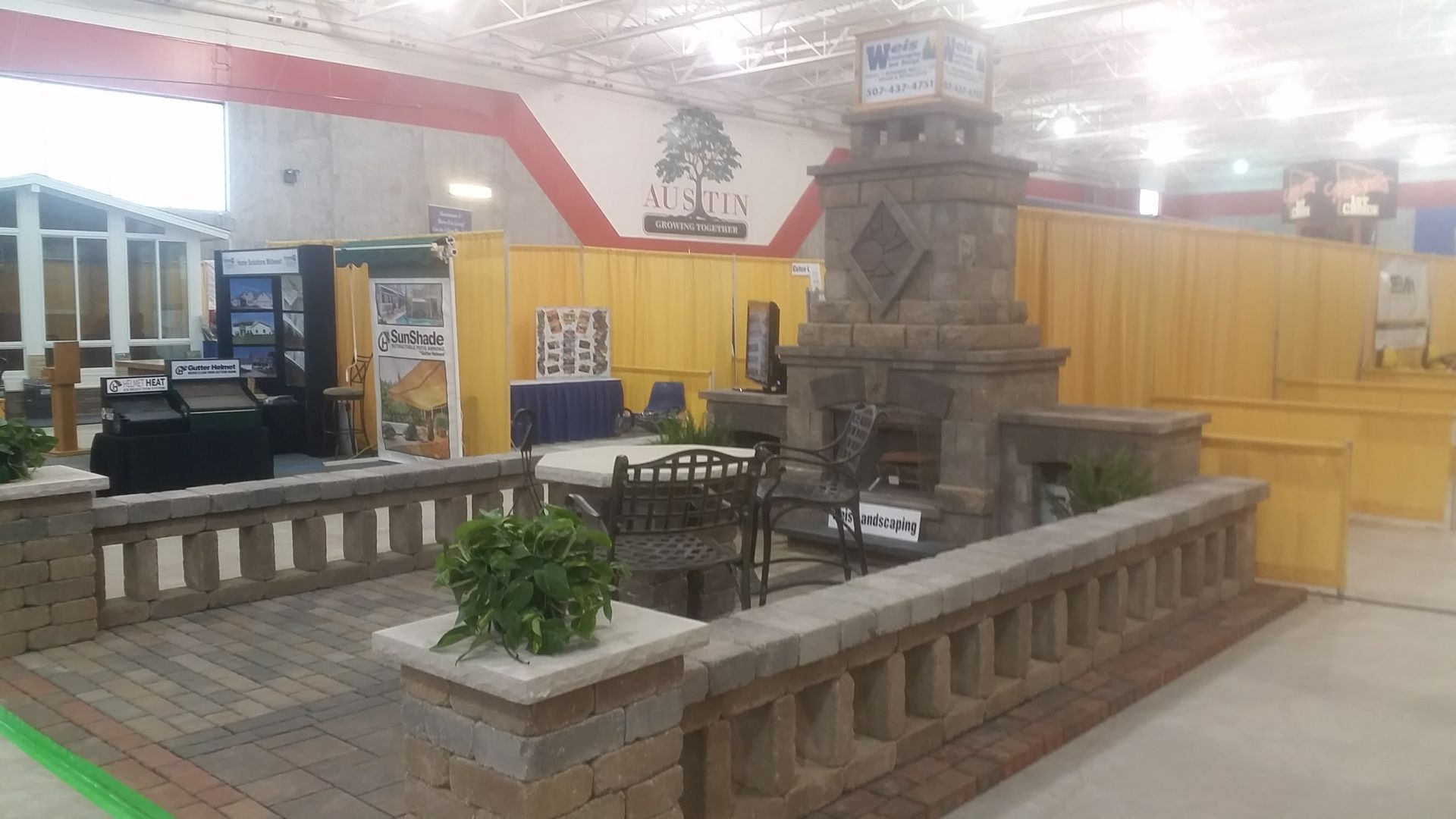 An indoor trade show display featuring a stone fireplace, paved patio area, and a stone-topped half-wall enclosure.