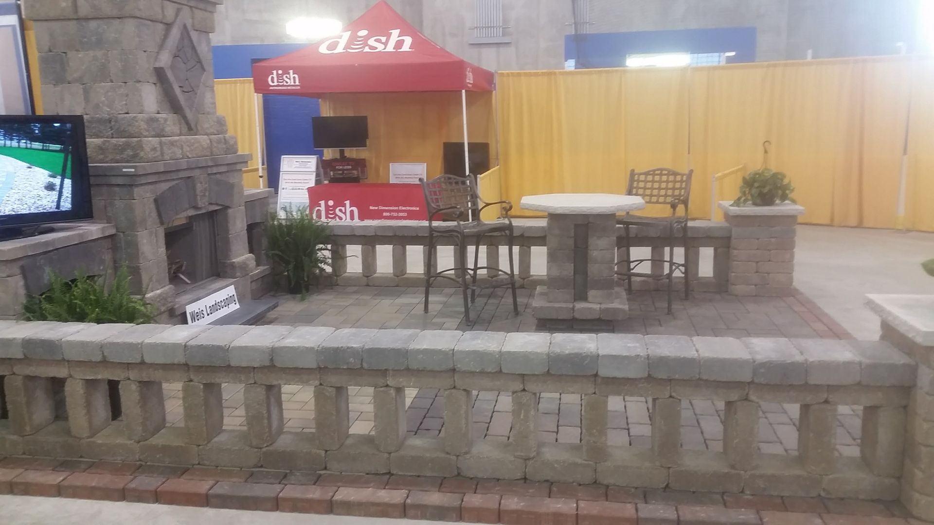 A stone patio display with a fireplace, a cafe table, chairs, and a red Dish Network tent in a convention hall.