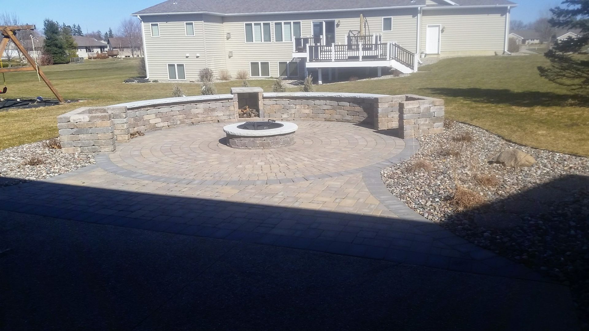A stone circular patio with a fire pit and a curved retaining wall in a grassy residential backyard.