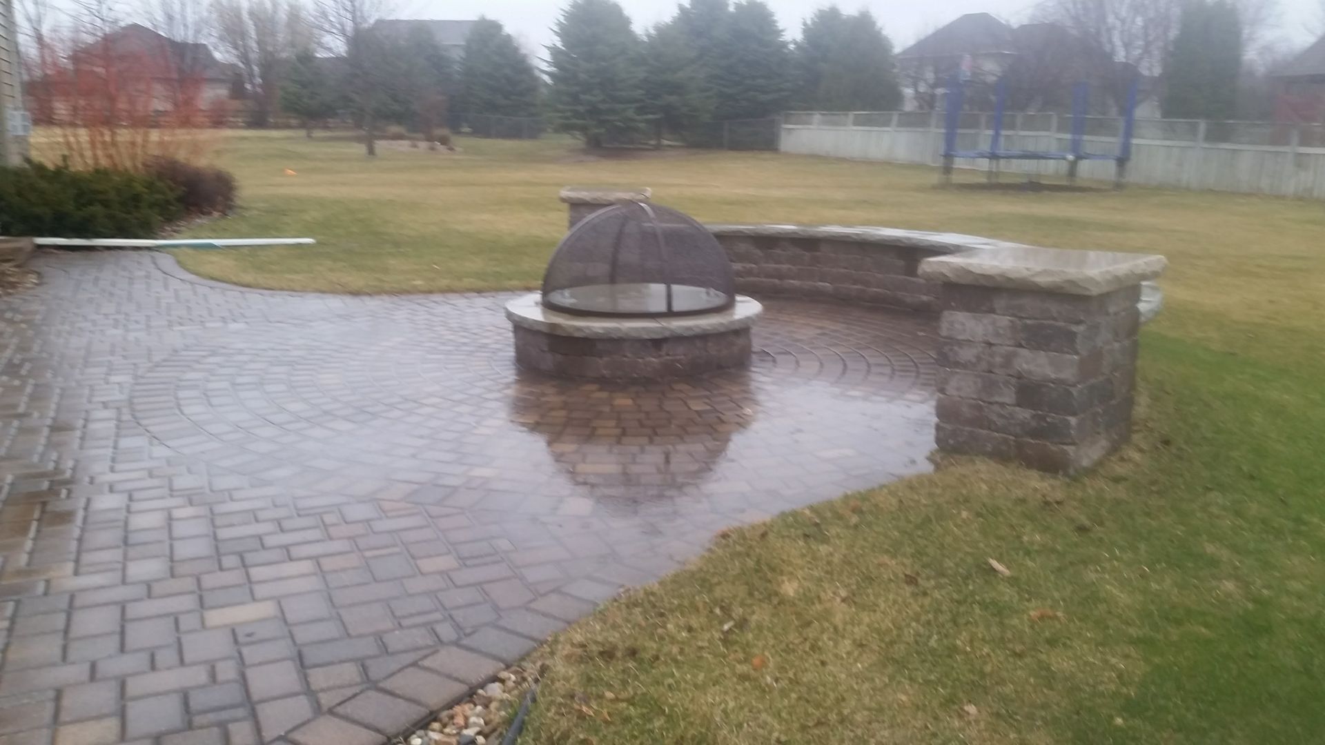 A stone patio with a fire pit and semi-circular seating wall in a residential backyard on an overcast day.