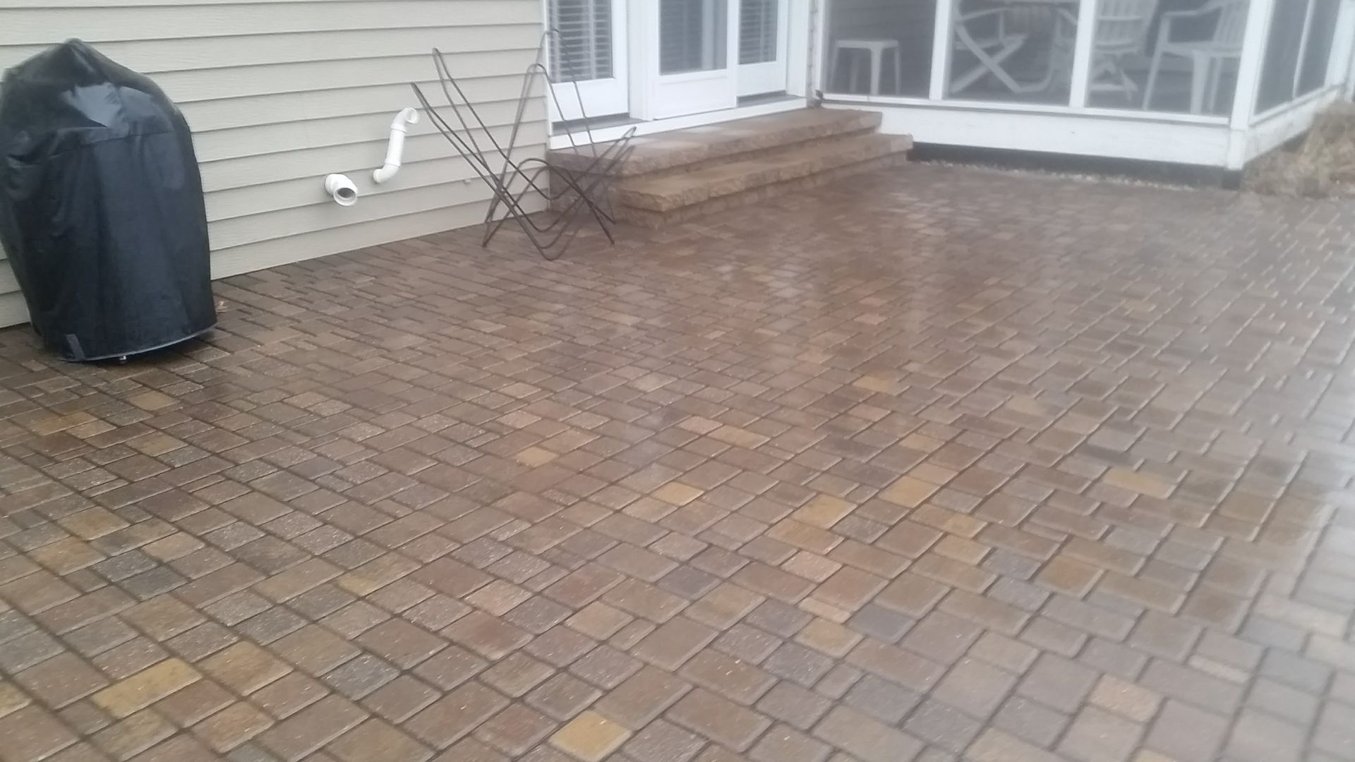 A paved stone patio with a grill in a black cover, located outside a house with a screen porch and white siding.